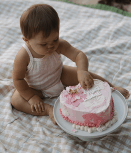 A baby sitting on a picnic blanket at the beach, wearing a light pink outfit and reaching into a small pink frosted cake on a white cake stand, with sand visible in the background.